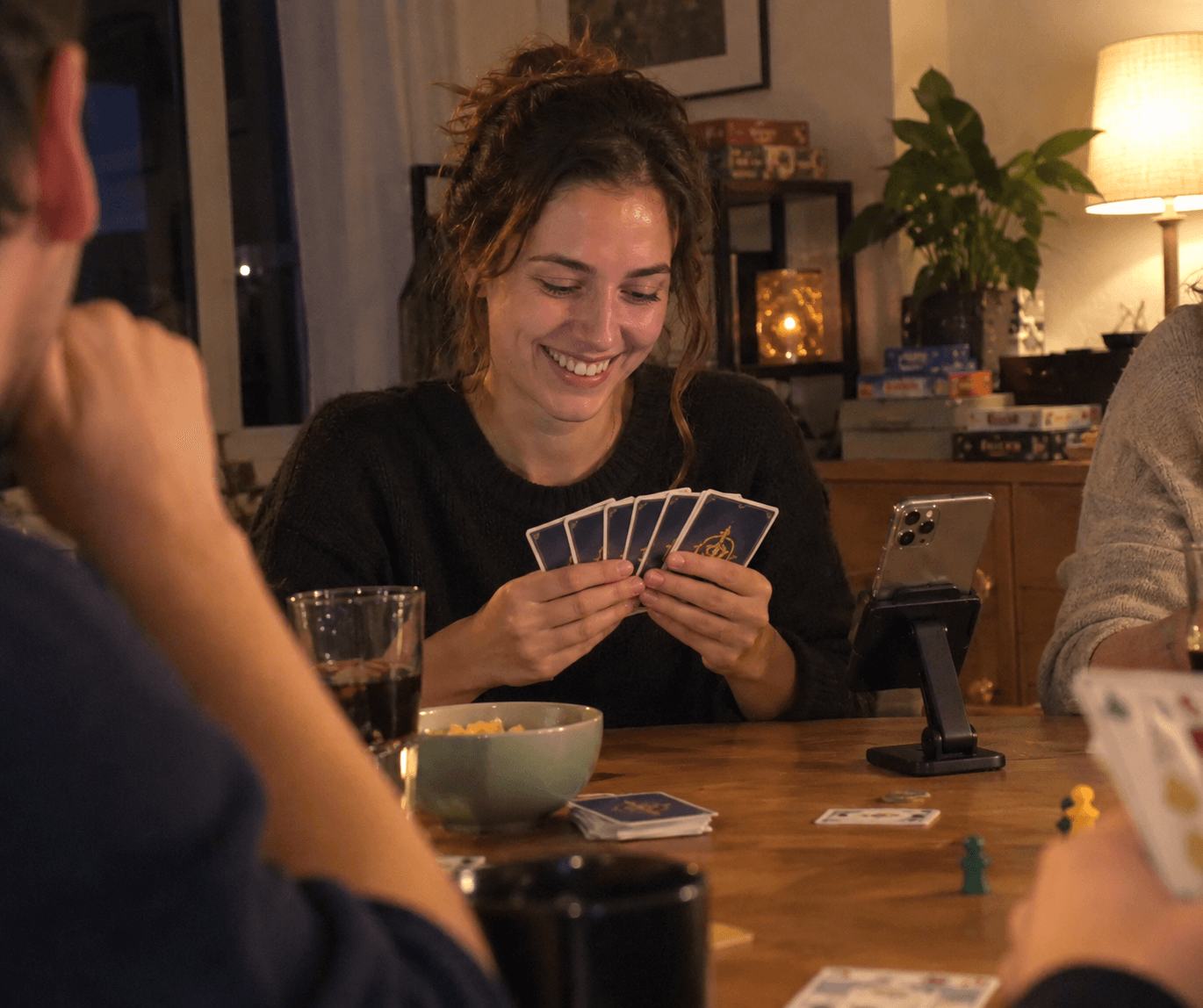 Game night at a wooden table: a woman laughs while holding her Crowns cards, next to her a phone with the app sits in a stand